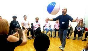 Teacher stands in circle of particpants throwing a large blue and while blow up ball